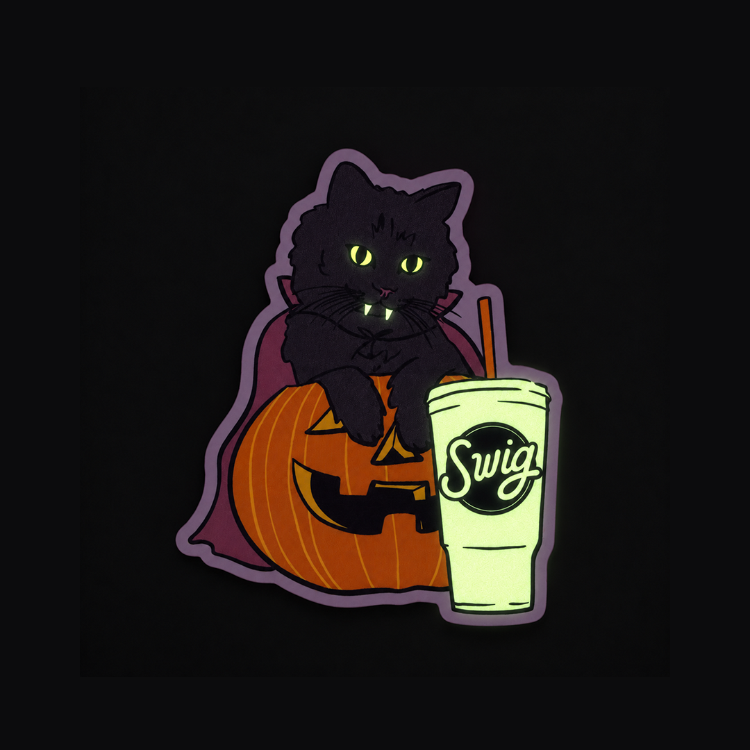 The glow in the dark cat sticker against darkness, with glowing green eyes, fangs and Swig cup beside an orange pumpkin.